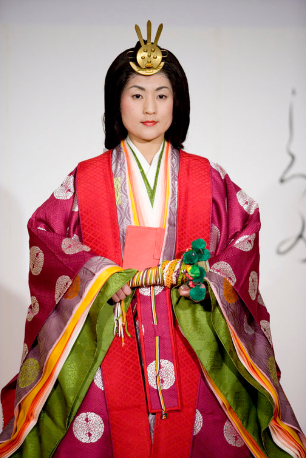 Eine Frau in einem traditionellen japanischen Kimono, das ein leuchtend rotes und grünes Kleid mit aufwändigen Mustern trägt, steht selbstbewusst auf einem Laufsteg, ihr Haar zu einem ordentlichen Knoten gebunden.