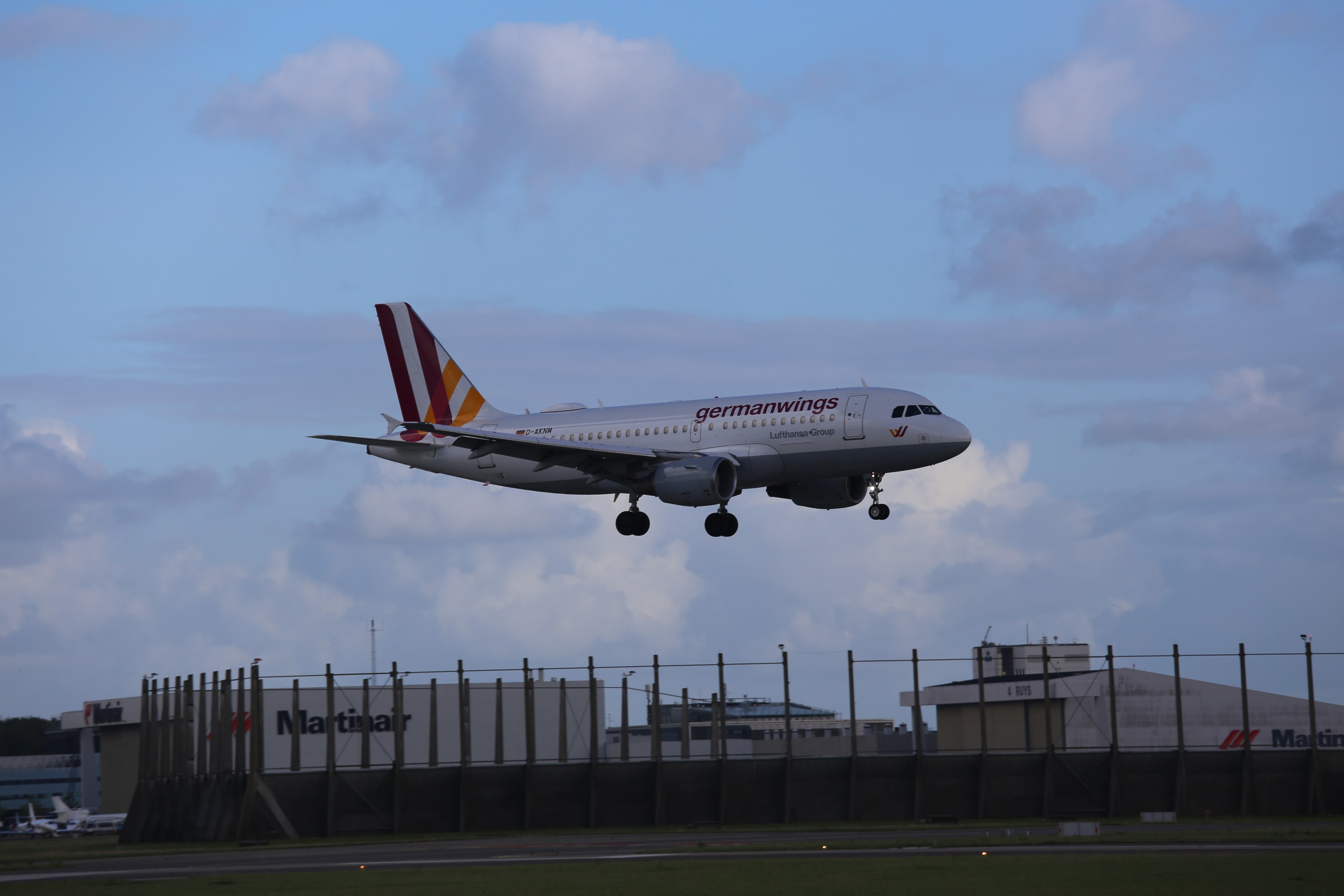 Germanwings Airbus A320-200 beim Abheben vom Frankfurt Airport mit Gras darunter, einem Zaun im Vordergrund, Gebäuden und Bäumen im Hintergrund und einer bewölkten Himmel
