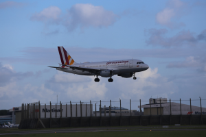 Germanwings Airbus A320-200 beim Abheben vom Frankfurt Airport mit Gras darunter, einem Zaun im Vordergrund, Gebäuden und Bäumen im Hintergrund und einer bewölkten Himmel