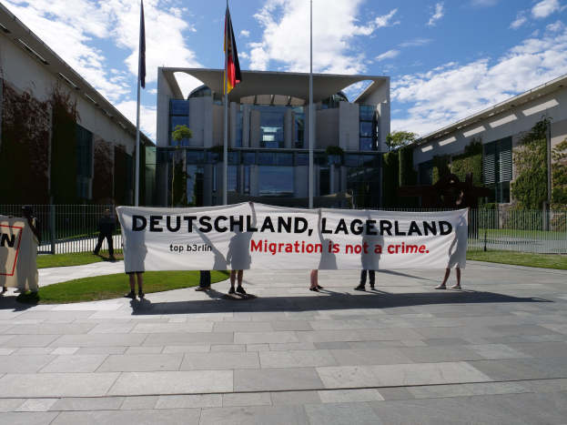 Eine Gruppe von Menschen hält ein Banner mit der Aufschrift "Deutschland, Lagerland Migration ist kein Verbrechen" vor einem umzäunten Gebäude mit Bäumen und Fahnen.