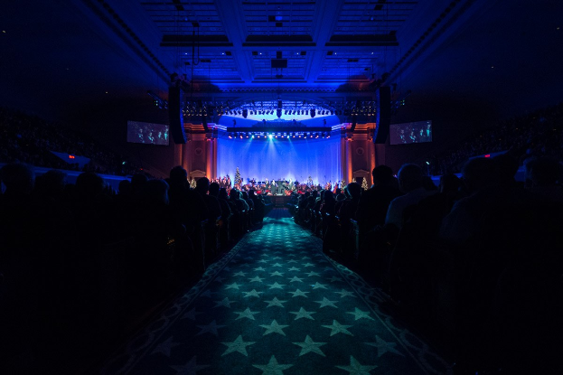 Ein großer Zuschauerraum mit Sitzplätzen, eine Bühne mit Musikern und eine Decke mit Beleuchtung und Fernsehgeräten, was auf ein Konzerthalle während der Nationalkonvention 2019 hindeutet.