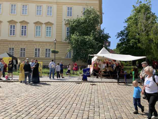 Eine Gruppe von Menschen geht eine Kopfsteinpflasterstraße in Krakau, Polen, entlang, vorbei an einem Gebäude mit Fenstern, Bäumen, Laternen und Zelten, wobei einige Mützen und Taschen tragen und Tische mit verschiedenen Gegenständen darauf stehen.