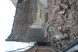 Teilweise abgerissener Bau mit verstreuten Ziegeln und Metallstangen, mit anderen Gebäuden und einem klaren blauen Himmel im Hintergrund.