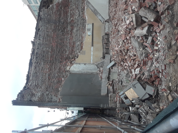 Teilweise abgerissener Bau mit verstreuten Ziegeln und Metallstangen, mit anderen Gebäuden und einem klaren blauen Himmel im Hintergrund.