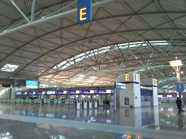 Leerer Flughafen-Terminal mit Menschen, die gehen, Schilder mit Text und Maschinen in der Mitte, eine Person links und Metallstangen und Lampen auf dem Dach.