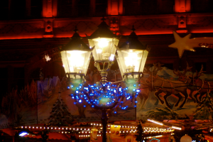 Eine Straßenlaterne in einer nächtlichen Stadt, geschmückt mit Lichtern und Dekorationen, steht vor einem Gebäude und verbreitet eine festliche Atmosphäre.