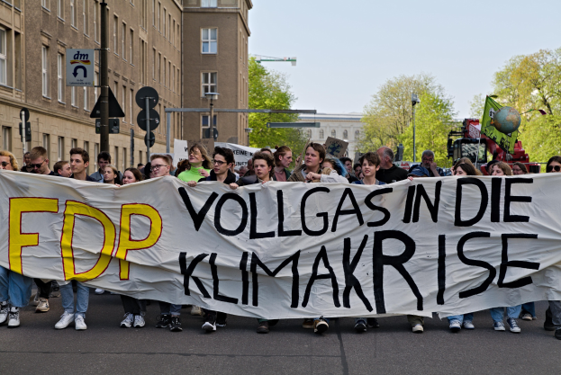 Gruppe von Menschen, die eine Straße entlanggehen und ein Banner mit der Aufschrift "FDP Vollgas in die Klimakrise" tragen, mit Gebäuden, Bäumen und Verkehrszeichen im Hintergrund unter einem klaren blauen Himmel.