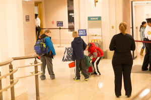 Eine Gruppe von Menschen steht in einem Flur mit einem Geländer auf der linken Seite, Deckenleuchten und Informationsschildern im Hintergrund, die für den ersten Schultag im U.S. Capitol beschriftet sind.
