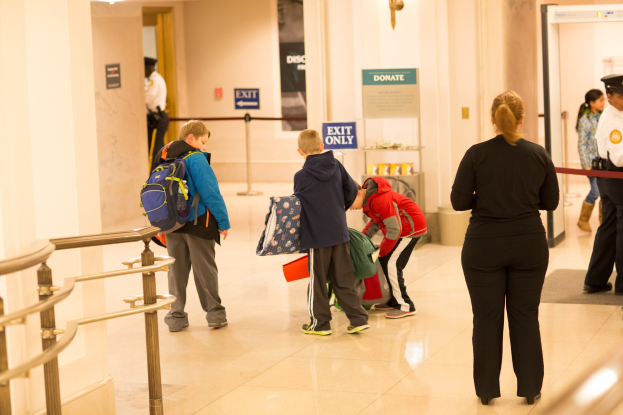 Eine Gruppe von Menschen steht in einem Flur mit einem Geländer auf der linken Seite, Deckenleuchten und Informationsschildern im Hintergrund, die für den ersten Schultag im U.S. Capitol beschriftet sind.