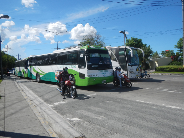 Ein grüner und weißer Shuttlebus steht am Straßenrand mit Motorradfahrern davor, ein grasbewachsener Fußweg links und Gebäude, Bäume, Laternenpfähle und einen klaren blauen Himmel im Hintergrund.