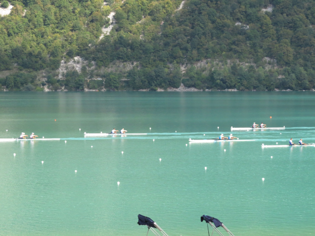 Gruppe von Menschen bei einem Ruderwettbewerb auf einem Gewässer mit Bäumen und einem Hügel im Hintergrund, Kleider über Ständen am Boden.