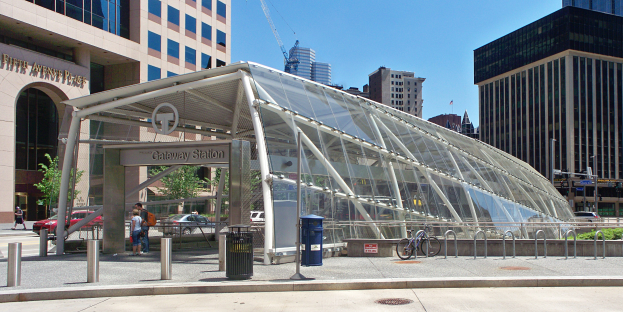 Eine belebte Stadtstraße mit hohen Wolkenkratzern, eine Bushaltestelle mit Glasdach, Menschen, Fahrzeuge, ein Metallzaun, ein Mülleimer, Pfosten, ein Schild, eine Verkehrsampel, Bäume, ein Kran und ein bewölkter Himmel.