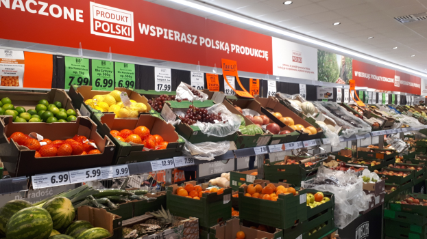 Ein Geschäftsraum mit verschiedenen Obst- und Gemüsesorten in Kartons mit Preisschildern, Informationsschilder an den Wänden und Deckenbeleuchtung.