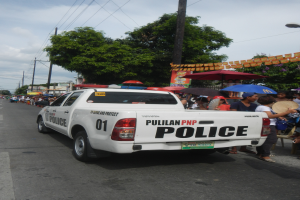 Ein Polizeiwagen fährt auf einer Straße mit Menschen am Straßenrand, einige halten Schirme, unter einem klaren blauen Himmel mit Bäumen, Polen, Drähten und Gebäuden im Hintergrund.