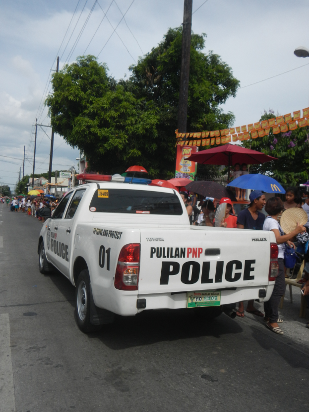 Ein Polizeiwagen fährt auf einer Straße mit Menschen am Straßenrand, einige halten Schirme, unter einem klaren blauen Himmel mit Bäumen, Polen, Drähten und Gebäuden im Hintergrund.