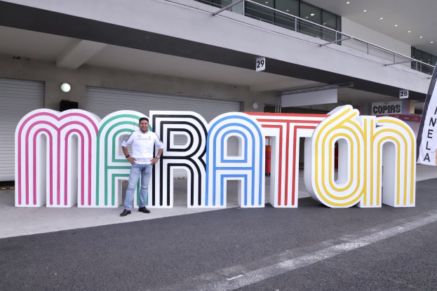 Ein Mann steht vor einem großen "Marathon"-Schild mit einem Banner rechts daneben, im Hintergrund ist ein Gebäude mit Lichtern, Geländern und Läden zu sehen.