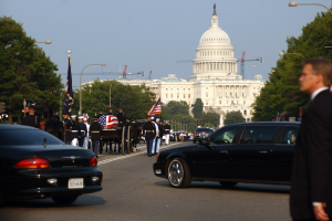 Eine Trauerprozession mit Menschen in Mützen und Fahnen vor dem Kapitolsgebäude, begleitet von Fahrzeugen, Laternenpfählen, Bäumen und Gebäuden unter einem klaren Himmel.