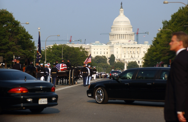 Eine Trauerprozession mit Menschen in Mützen und Fahnen vor dem Kapitolsgebäude, begleitet von Fahrzeugen, Laternenpfählen, Bäumen und Gebäuden unter einem klaren Himmel.